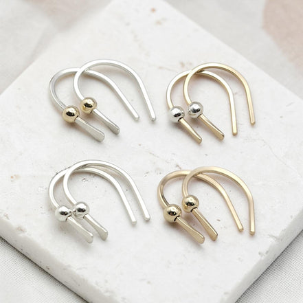 Four minimalist U-shaped hoop earrings with small metallic ball accents, shown in silver and gold finishes arranged on a white marble slab.