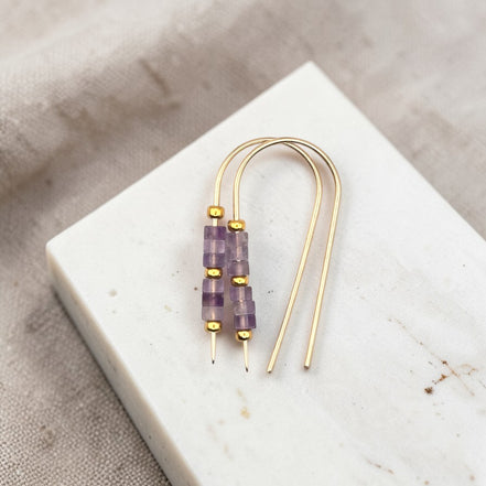 Gold earrings with purple amethyst beads on a white stone surface