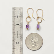 Gold earrings with purple beads next to a ruler and quarter for scale on a white background