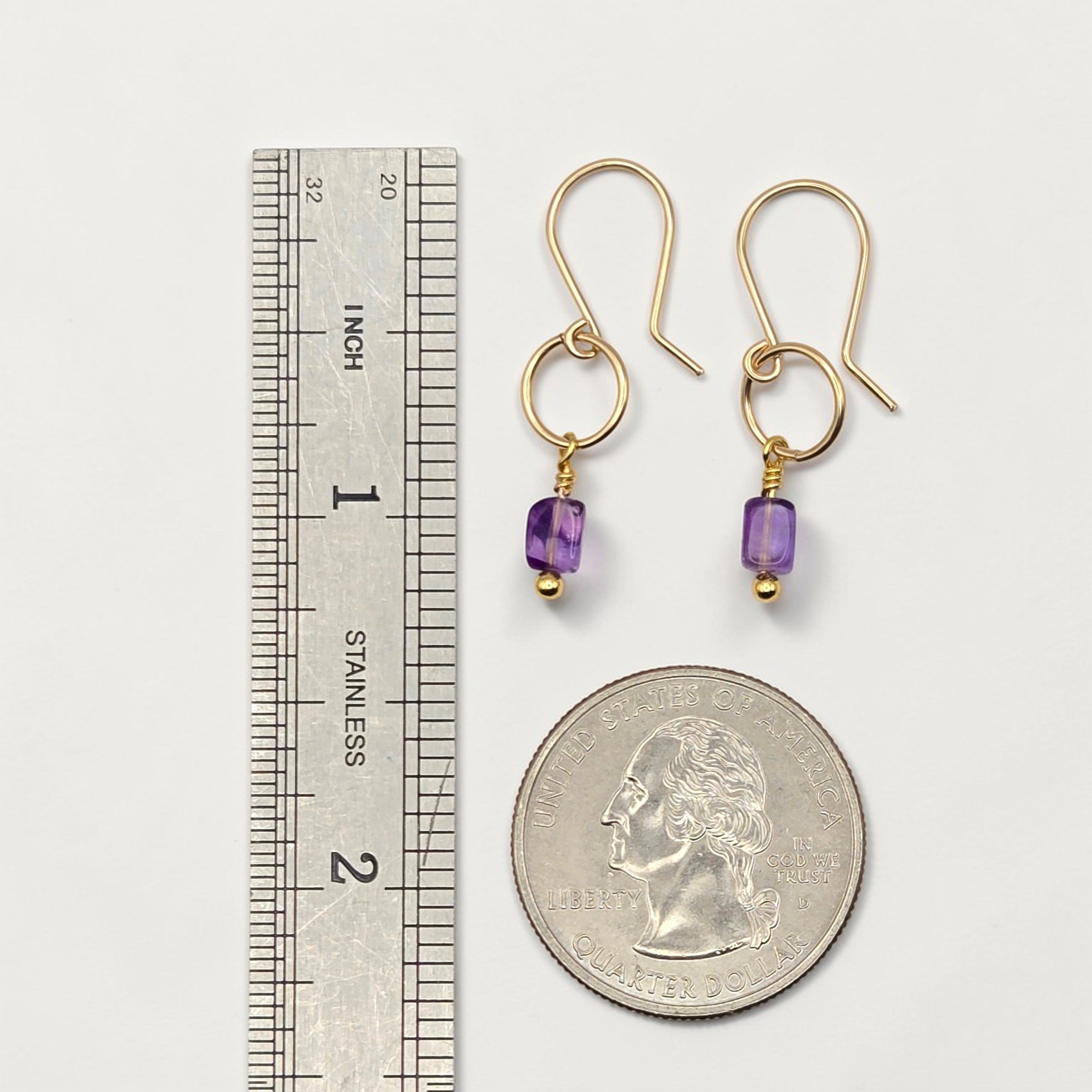 Gold earrings with purple beads next to a ruler and quarter for scale on a white background