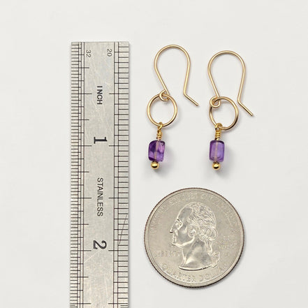 Gold earrings with purple beads next to a ruler and quarter for scale on a white background