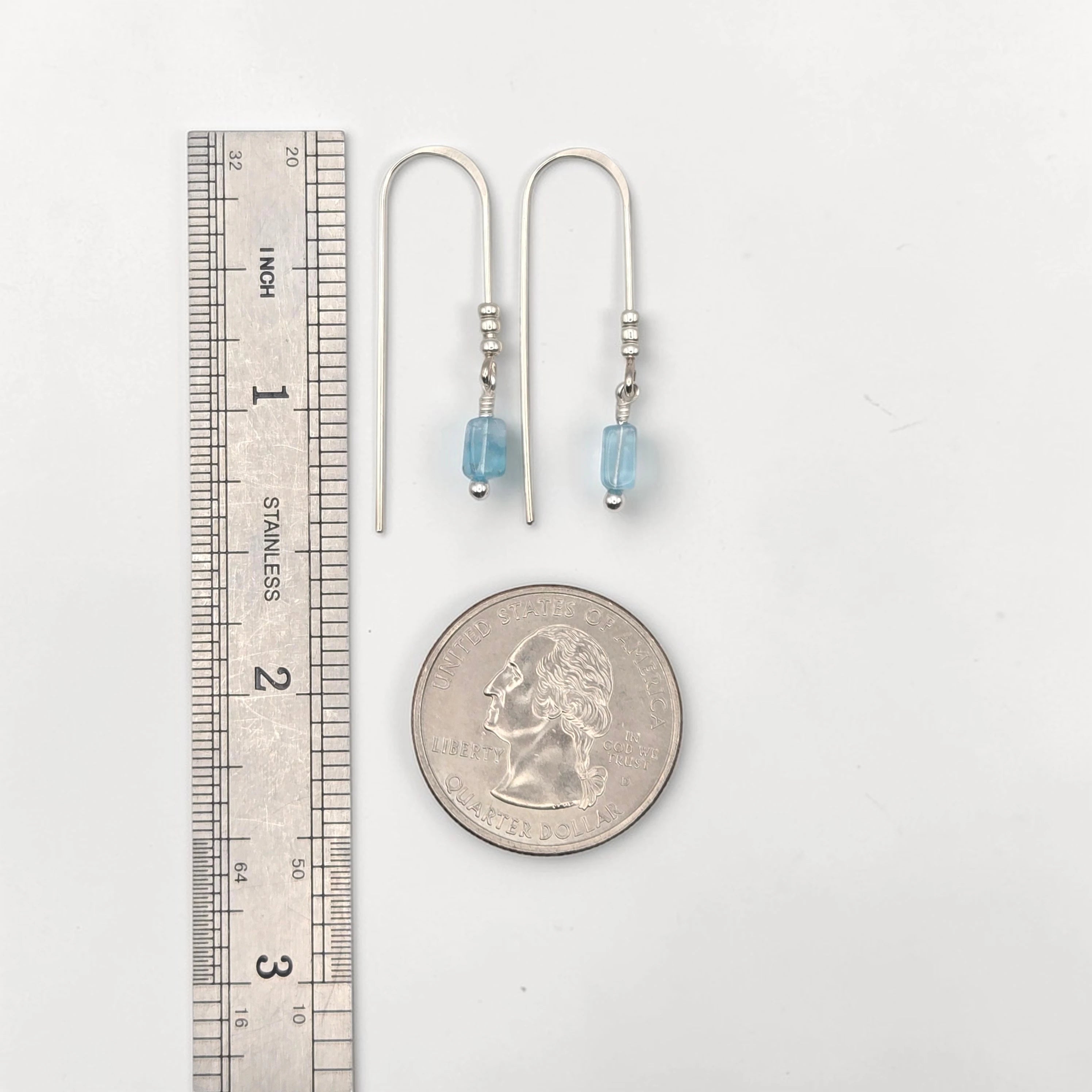 Pair of silver earrings with blue beads next to a ruler and quarter for scale on a white background