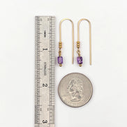 Gold earrings with purple beads next to a ruler and quarter for scale on a white background
