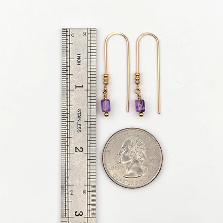 Gold earrings with purple beads next to a ruler and quarter for scale on a white background