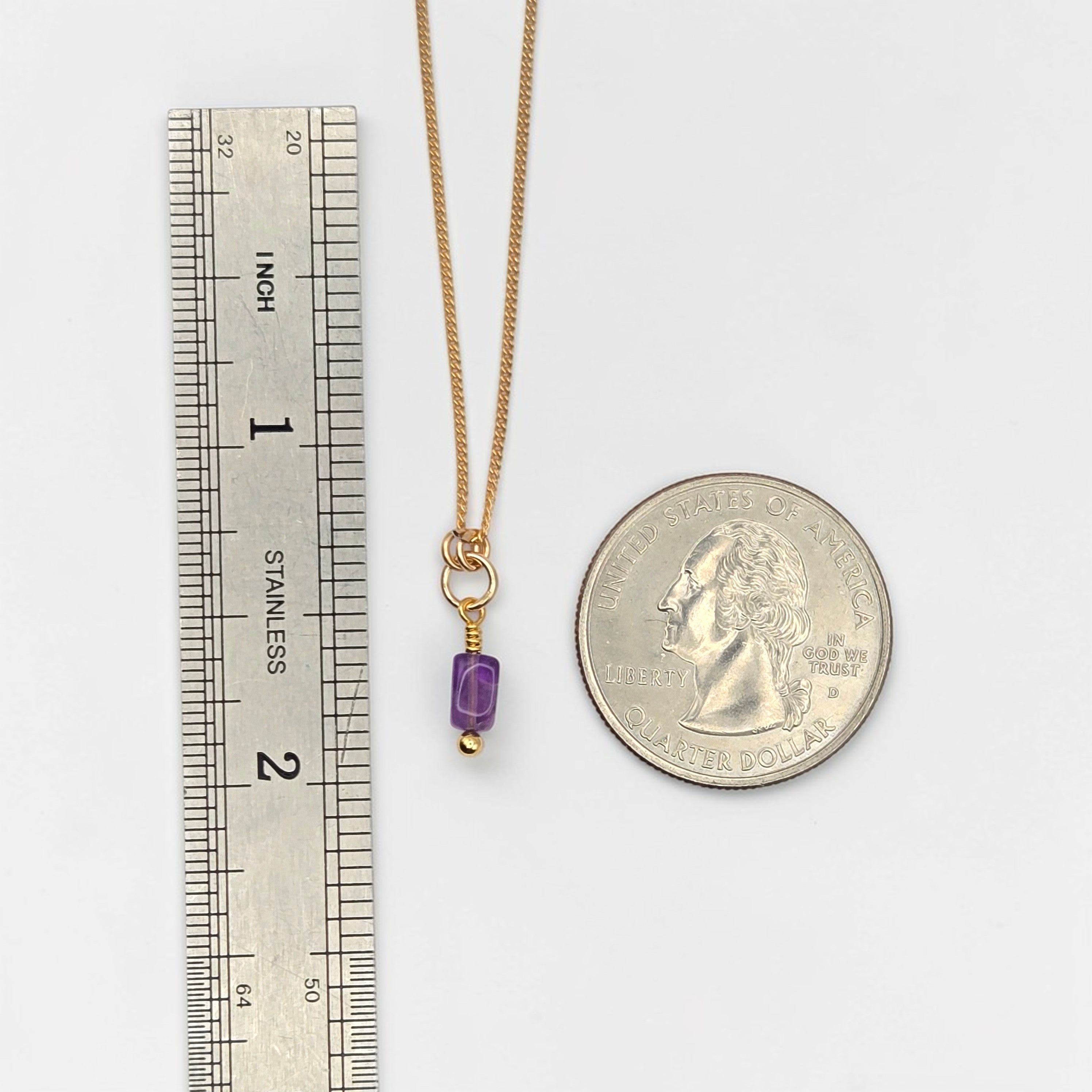 Gold necklace with a purple pendant next to a ruler and a coin for scale on a white background