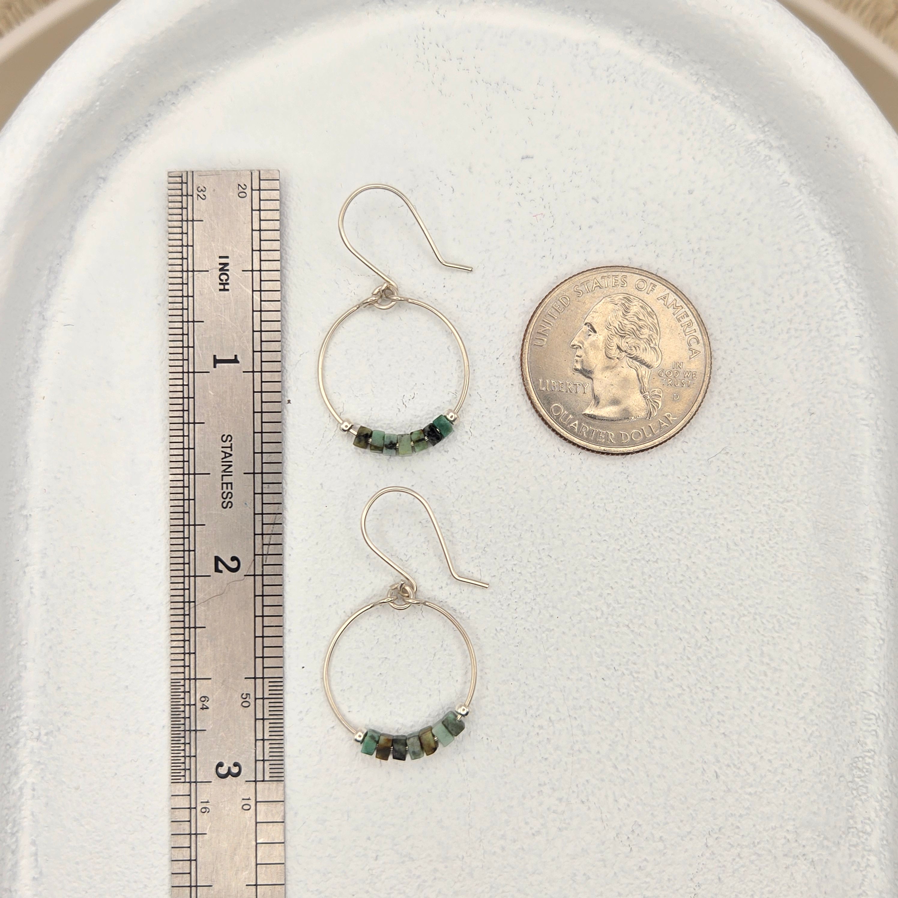 Two hoop earrings with green beads on a white surface with a coin and ruler for scale.