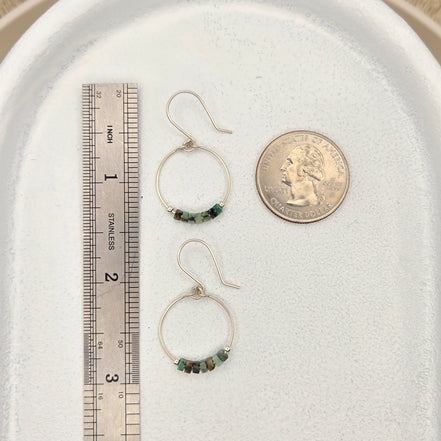 Two hoop earrings with green beads on a white surface with a coin and ruler for scale.