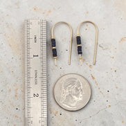 Gold earrings with black beads next to a ruler and quarter for scale on a textured surface.