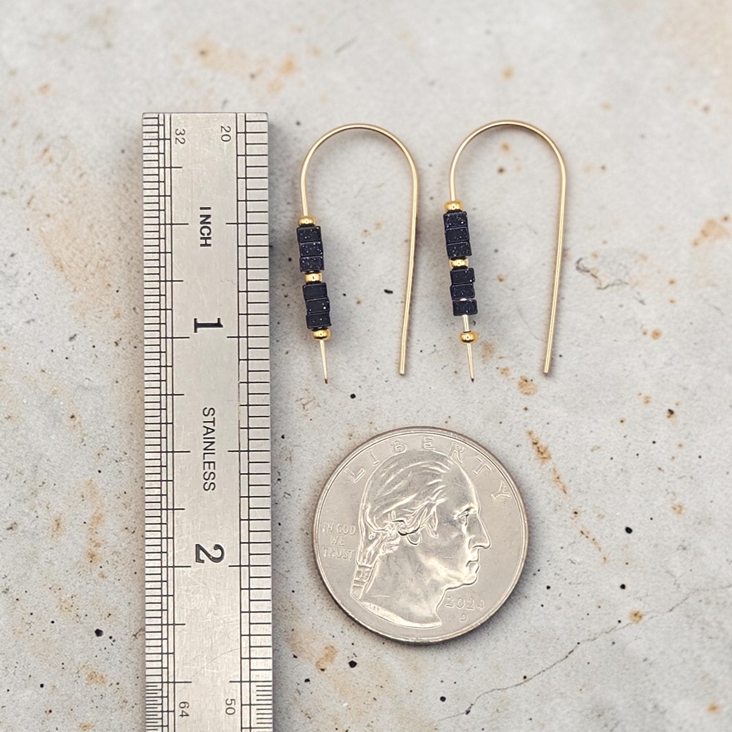 Gold earrings with black beads next to a ruler and quarter for scale on a textured surface.