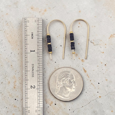 Gold earrings with black beads next to a ruler and quarter for scale on a textured surface.