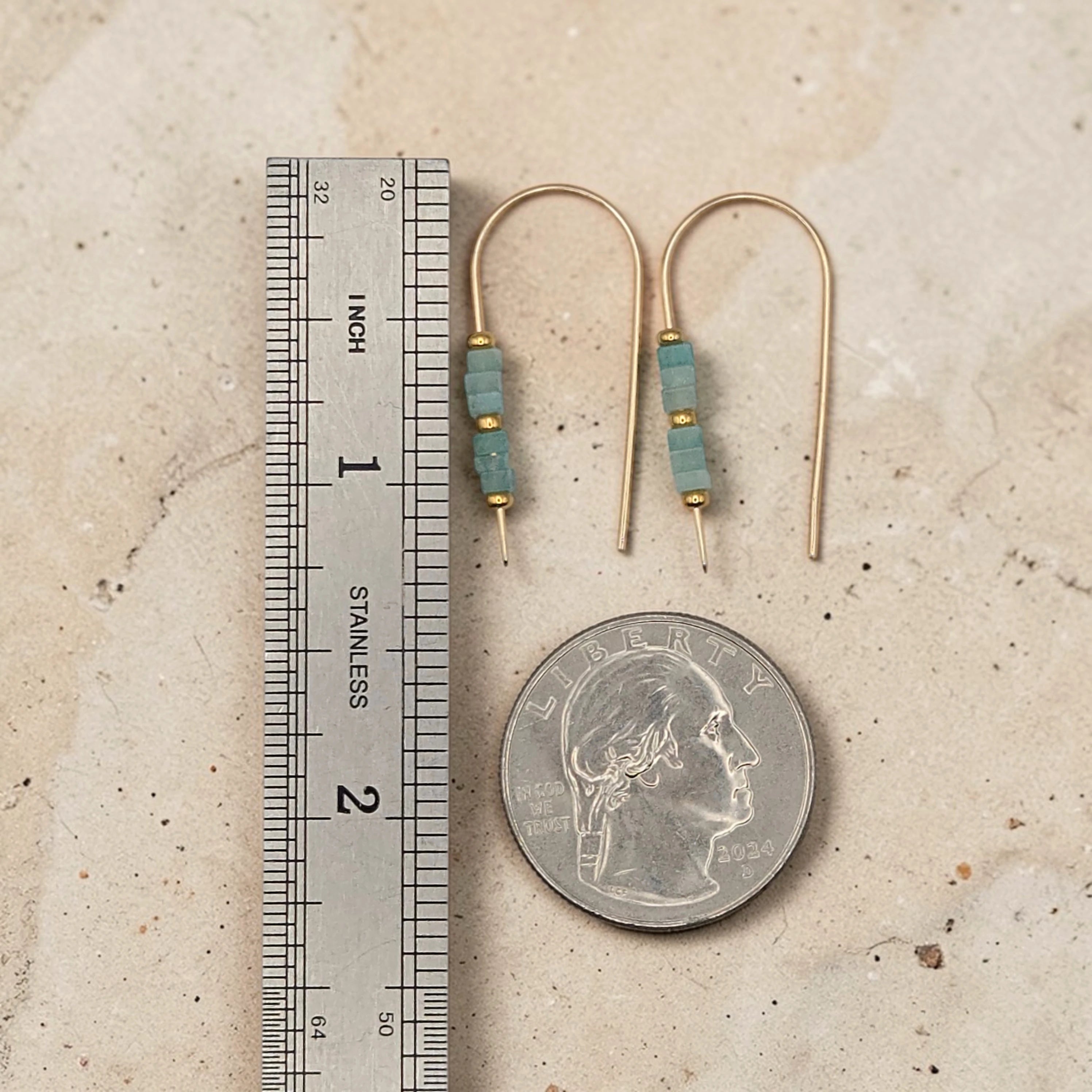 Gold earrings with blue beads next to a ruler and quarter for scale on a beige surface
