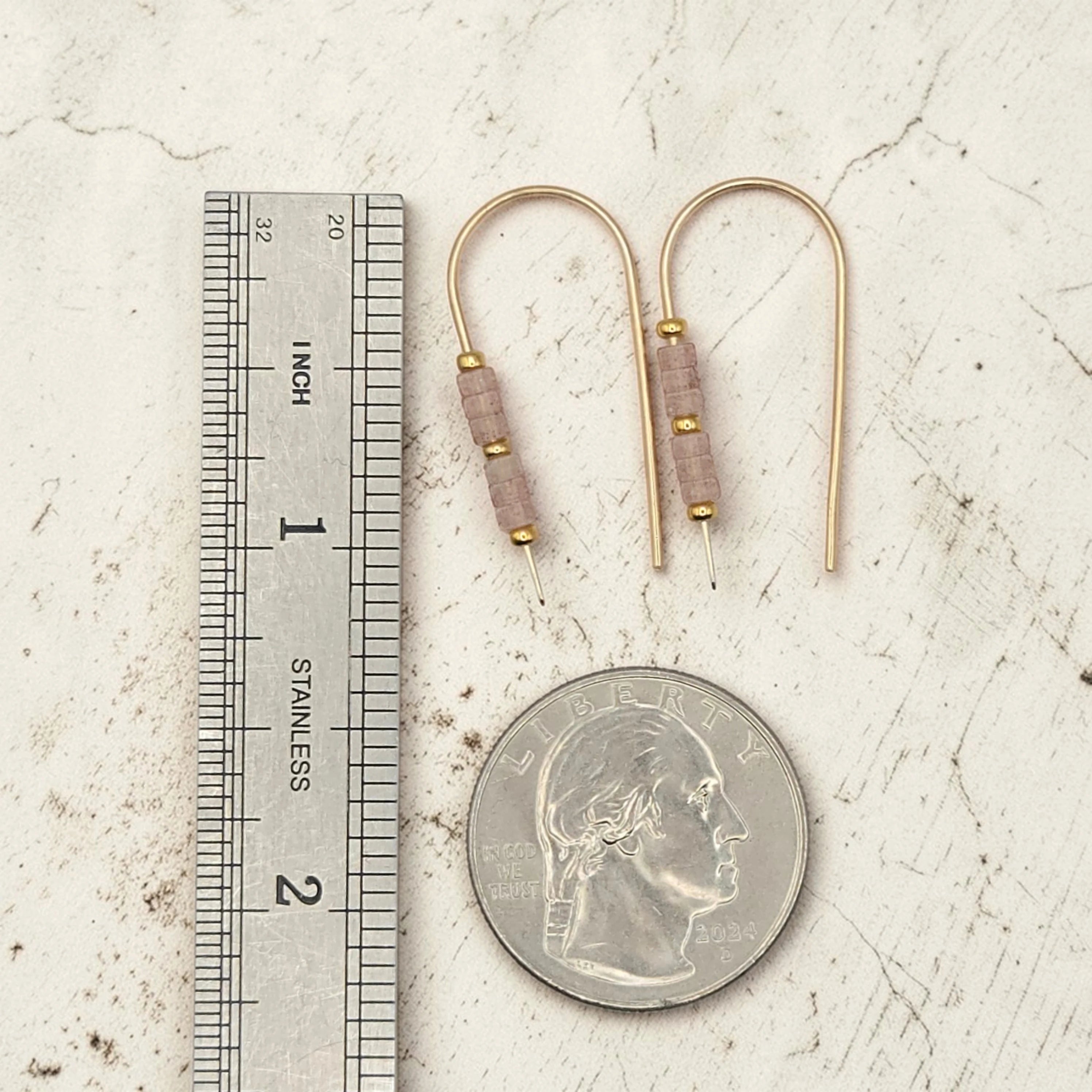 Gold earrings with brown beads next to a ruler and quarter for scale on a textured surface
