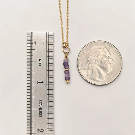 Gold necklace with purple beads next to a ruler and a quarter for scale on a white background