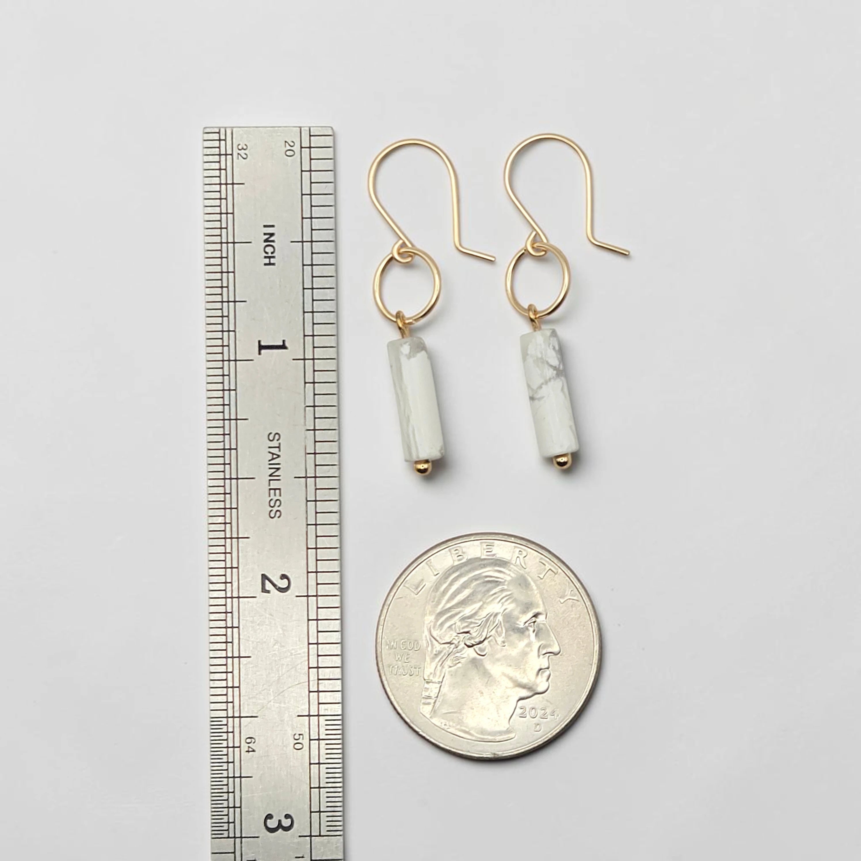Gold earrings with cylindrical beads next to a ruler and quarter for scale on a light gray background
