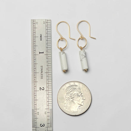 Gold earrings with cylindrical beads next to a ruler and quarter for scale on a light gray background