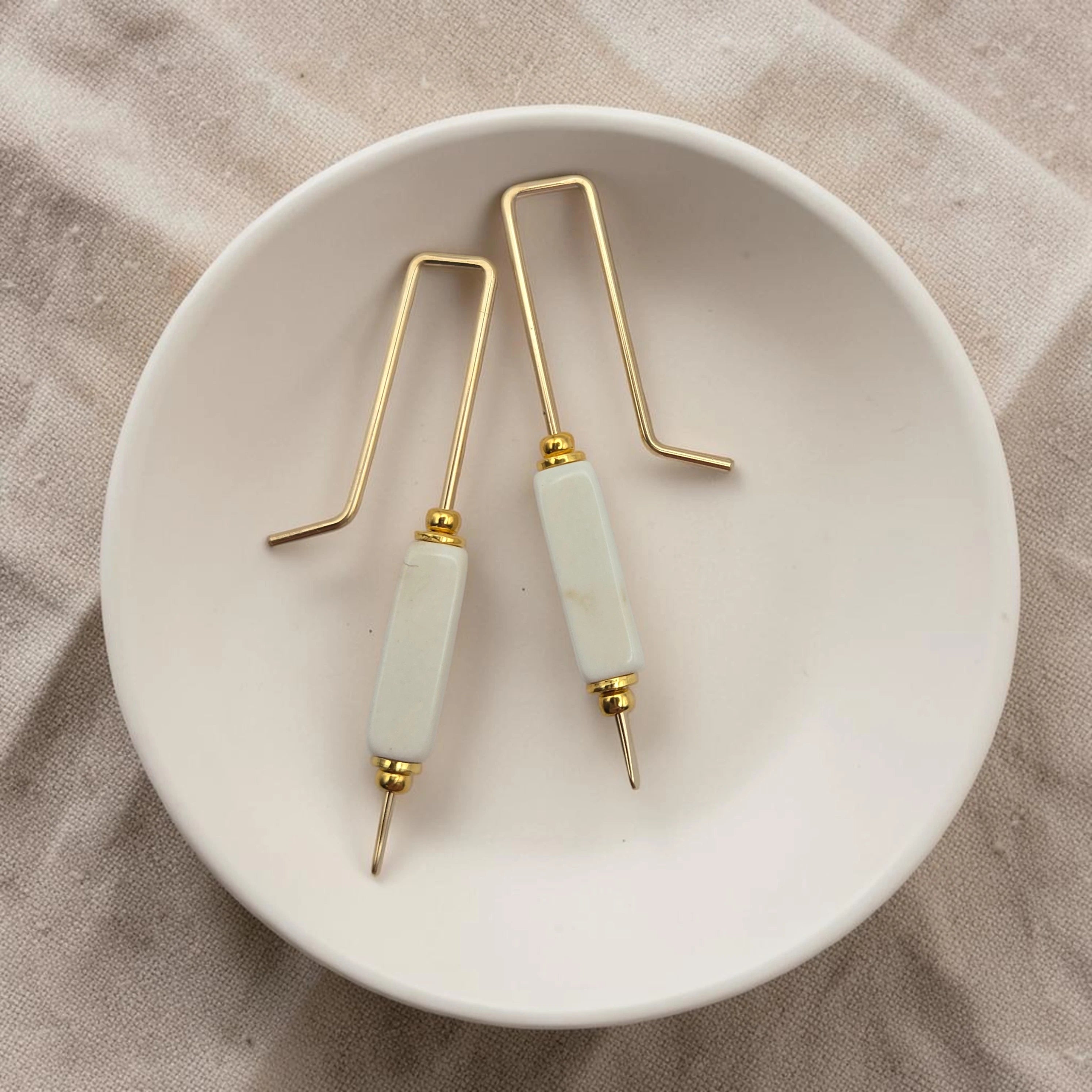 Pair of gold and light cream earrings on a white dish with a beige fabric background