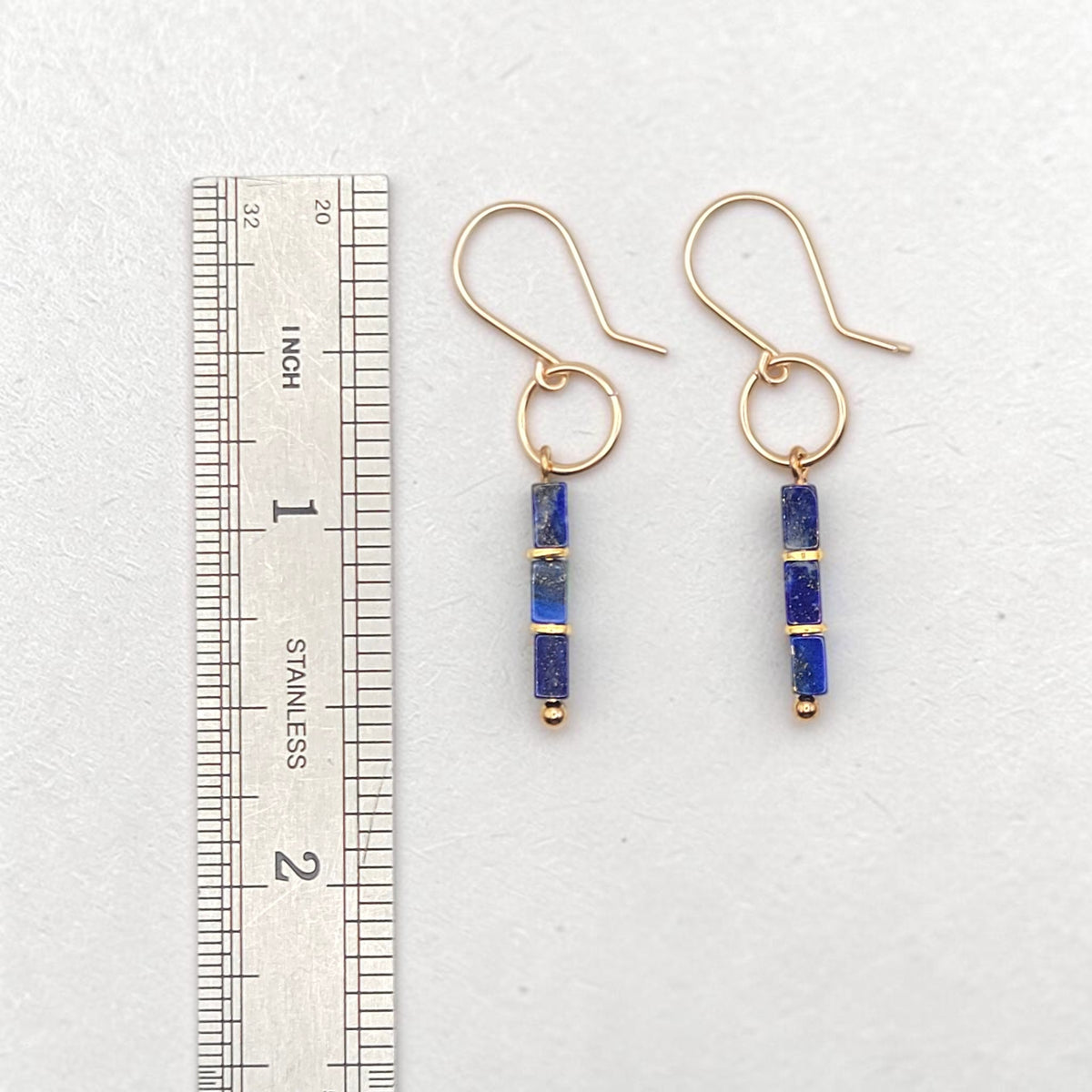 A pair of gold dangle earrings with blue lapis lazuli cube beads, shown next to a stainless steel ruler for scale on a light gray background.