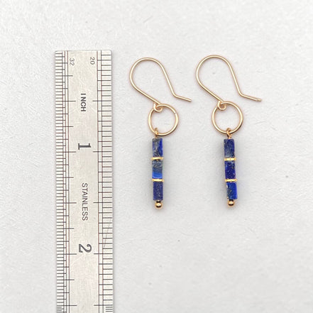 A pair of gold dangle earrings with blue lapis lazuli cube beads, shown next to a stainless steel ruler for scale on a light gray background.
