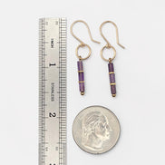 A pair of gold drop earrings with purple agate beads, shown next to a stainless steel ruler and a US quarter coin for scale, on a light gray background.