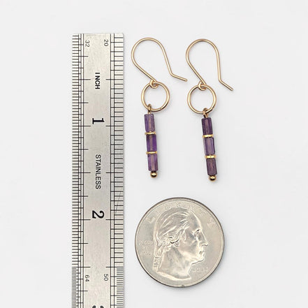 A pair of gold drop earrings with purple agate beads, shown next to a stainless steel ruler and a US quarter coin for scale, on a light gray background.