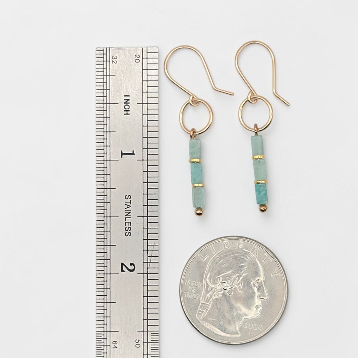 A pair of gold drop earrings with light blue turquoise beads and small gold spacer rings, shown next to a stainless steel ruler and a US quarter for scale on a white background.