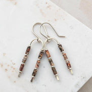 A pair of handmade dangle earrings with silver-toned fishhook wires and cylindrical beads featuring a leopard skin jasper that is natural brown, gray, and black marbled pattern, displayed on a light marble surface.