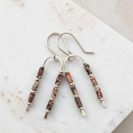 A pair of handmade dangle earrings with silver-toned fishhook wires and cylindrical beads featuring a leopard skin jasper that is natural brown, gray, and black marbled pattern, displayed on a light marble surface.