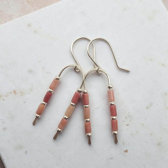 A pair of silver-toned dangle earrings with multiple small rectangular beads in soft pink, peach, and coral tones, resting on a light marble surface.