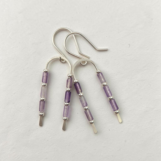 Three silver dangle earrings with amethyst beads, displayed on a textured off-white background.