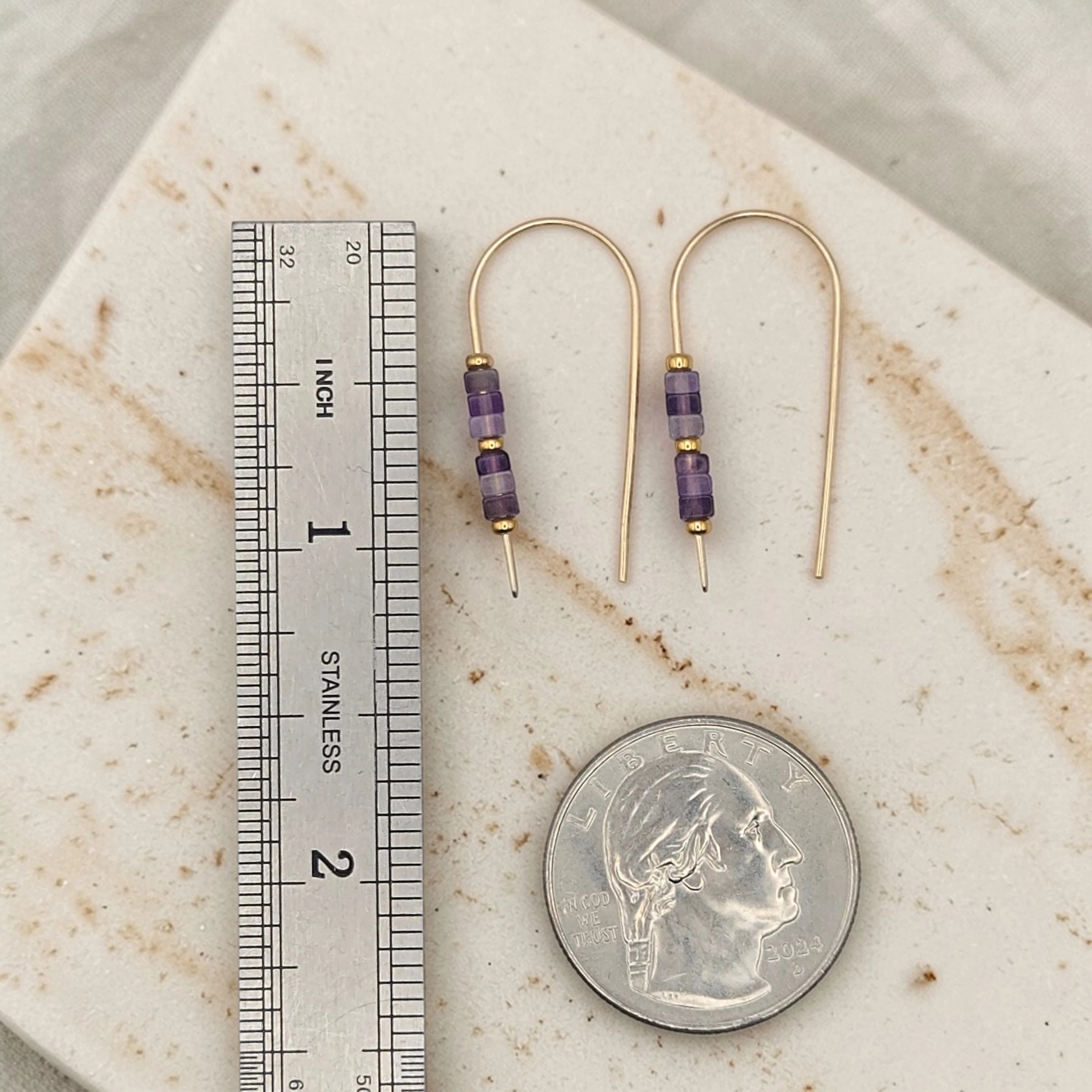 Gold earrings with purple beads next to a ruler and quarter for scale on a marble surface