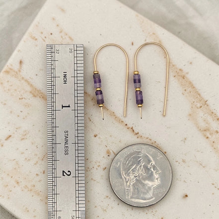 Gold earrings with purple beads next to a ruler and quarter for scale on a marble surface