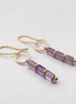 A pair of gold drop earrings with three faceted amethyst beads on each earring, displayed on a clean, light gray textured surface.