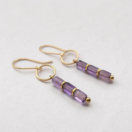 A pair of gold drop earrings with three faceted amethyst beads on each earring, displayed on a clean, light gray textured surface.