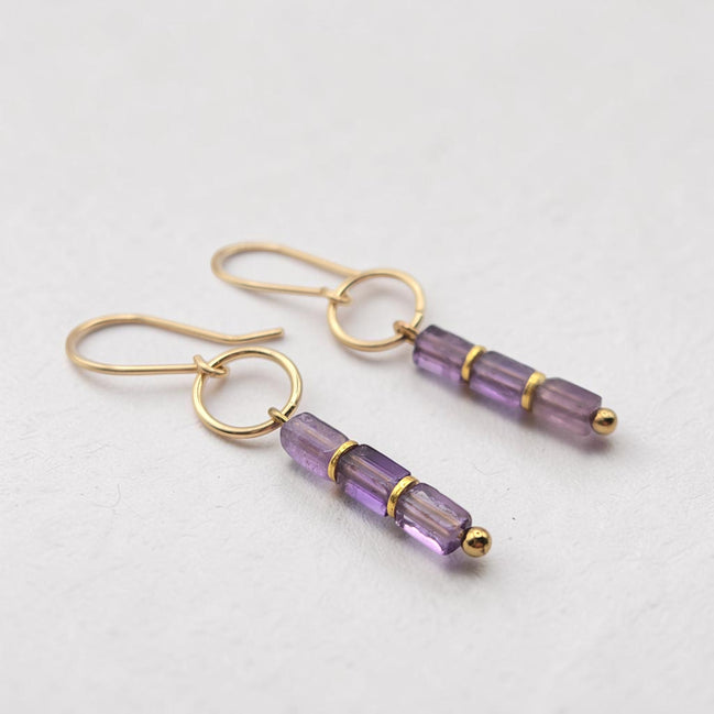 A pair of gold drop earrings with three faceted amethyst beads on each earring, displayed on a clean, light gray textured surface.