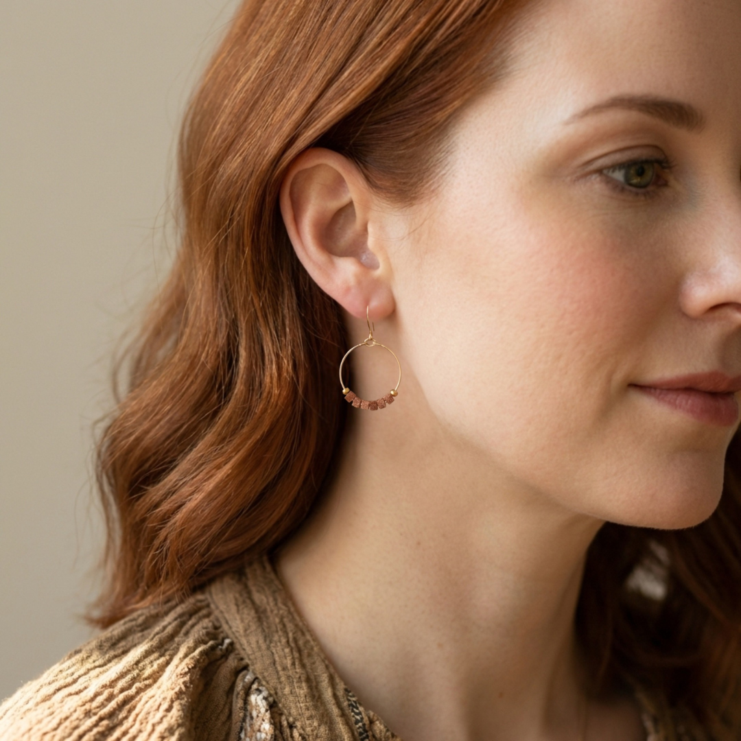 Close-up of a woman wearing gold hoop earrings with red gemstones against a neutral background