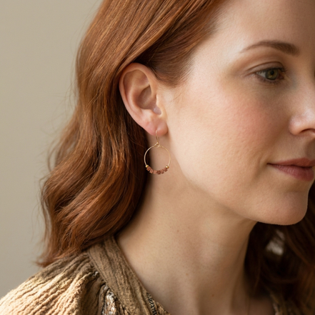 Close-up of a woman wearing gold hoop earrings with red gemstones against a neutral background