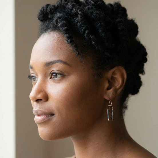 A close-up side profile of a woman with short, curly black hair wearing a delicate gold hoop earring with two small blue bead drops, set against a soft beige background.