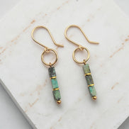 A pair of gold dangle earrings with rectangular green and gray stone beads, displayed on a white marble slab.