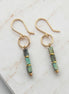 A pair of gold dangle earrings with rectangular green and gray stone beads, displayed on a white marble slab.