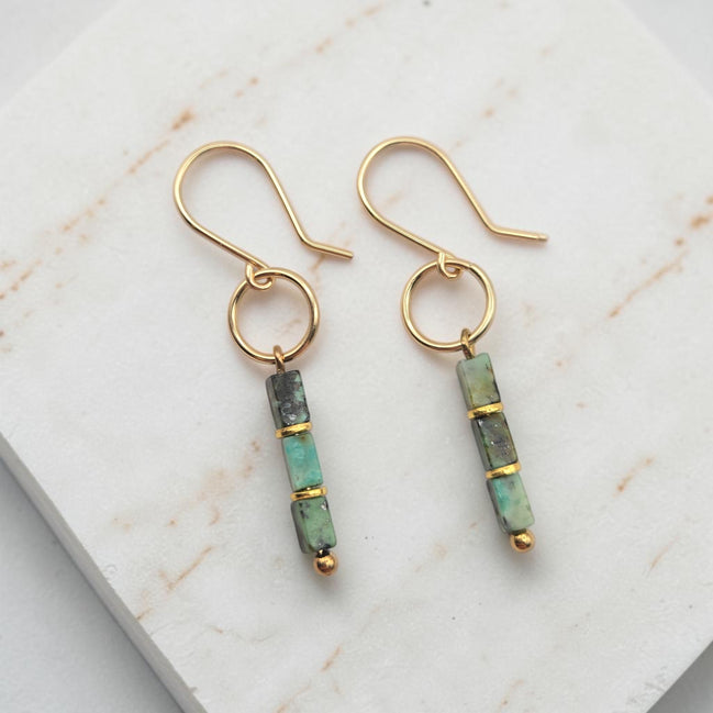 A pair of gold dangle earrings with rectangular green and gray stone beads, displayed on a white marble slab.