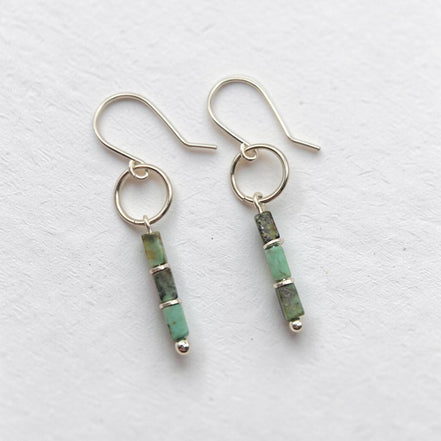 A pair of silver dangle earrings with small green and black african turquoise bead drops, displayed on a textured white surface.