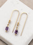 Gold earrings with purple amethyst beads on a white jewelry display box.