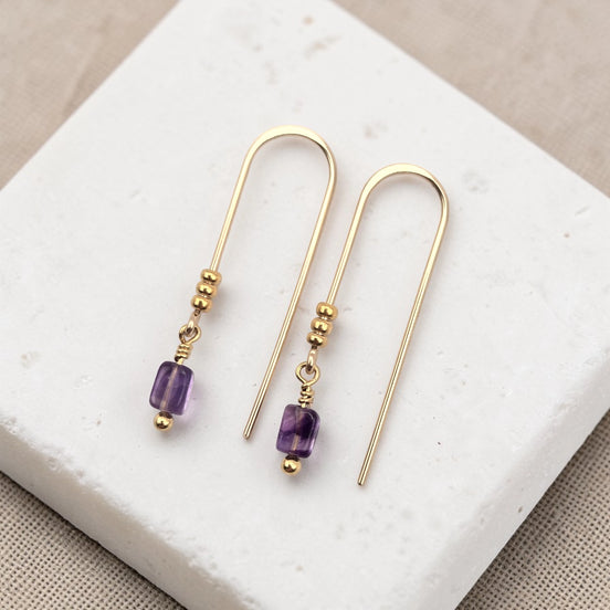 Gold earrings with purple amethyst beads on a white jewelry display box.