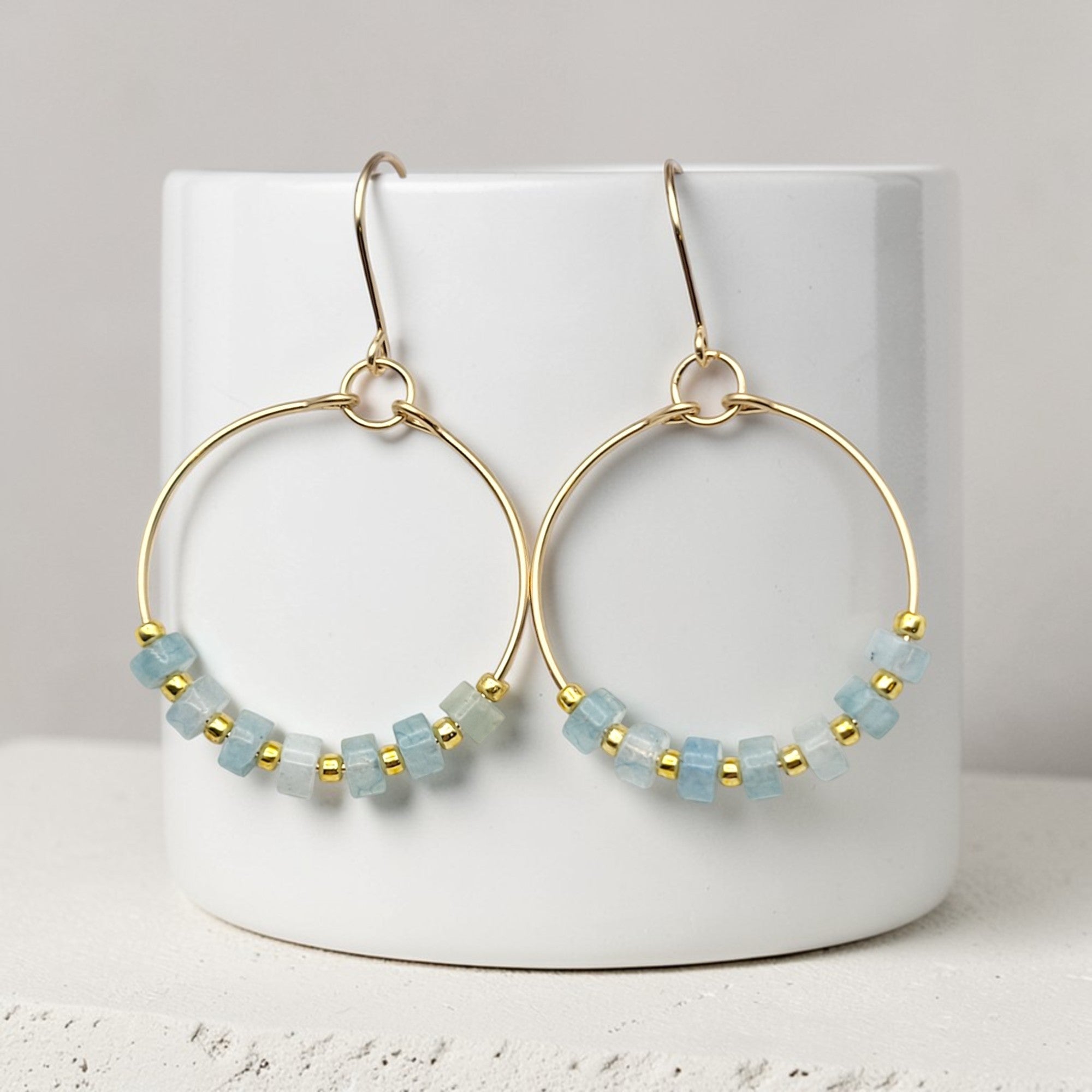 Gold hoop earrings with light blue beads on a white cylindrical container.