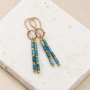 Bijou by SAM Still Water dangle earrings featuring blue apatite tube beads and 14k gold fill hoops on stone surface.