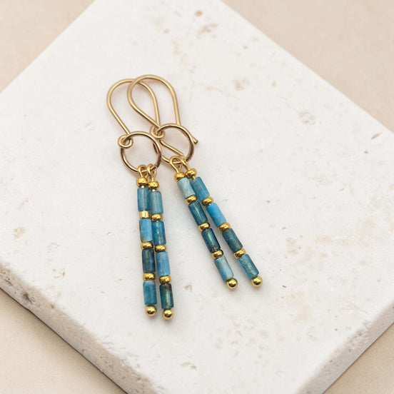 Bijou by SAM Still Water dangle earrings featuring blue apatite tube beads and 14k gold fill hoops on stone surface.
