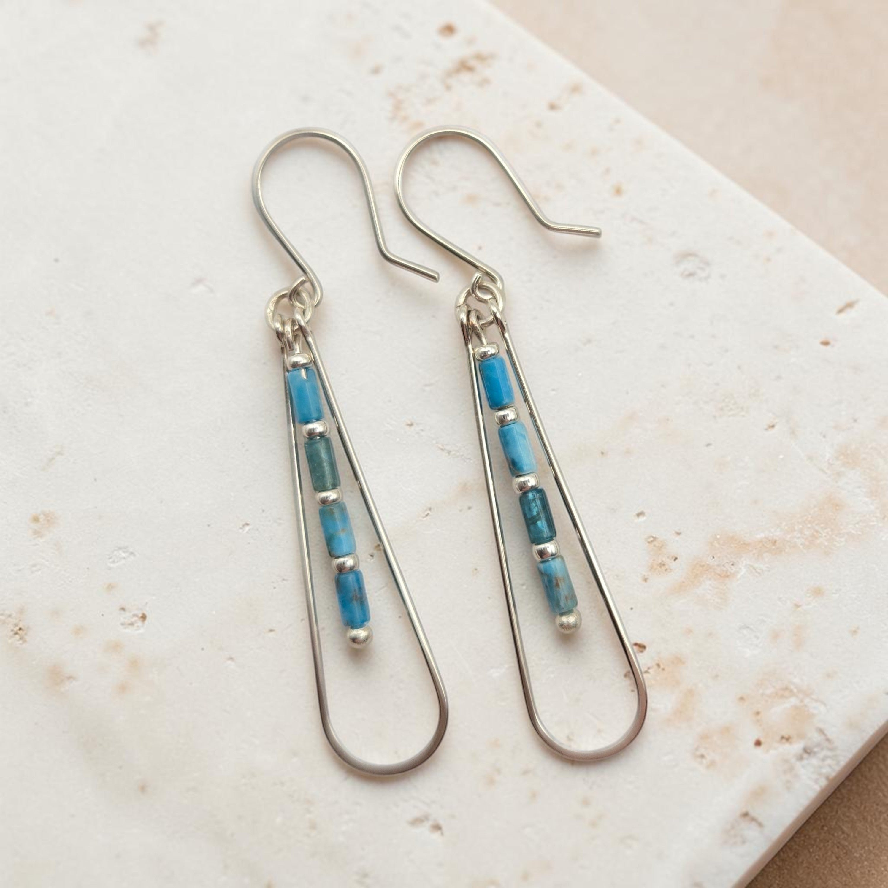Bijou by SAM Still Water teardrop earrings with blue Apatite tube beads and sterling silver hoops on stone surface.