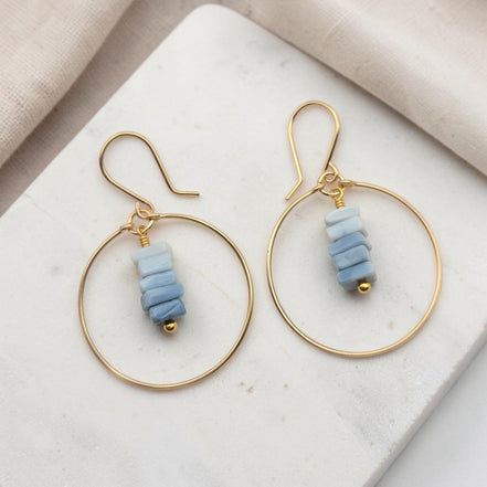 Bijou by SAM gold hoop earrings featuring a delicate stack of square-cut denim blue opal stones on a marble surface.