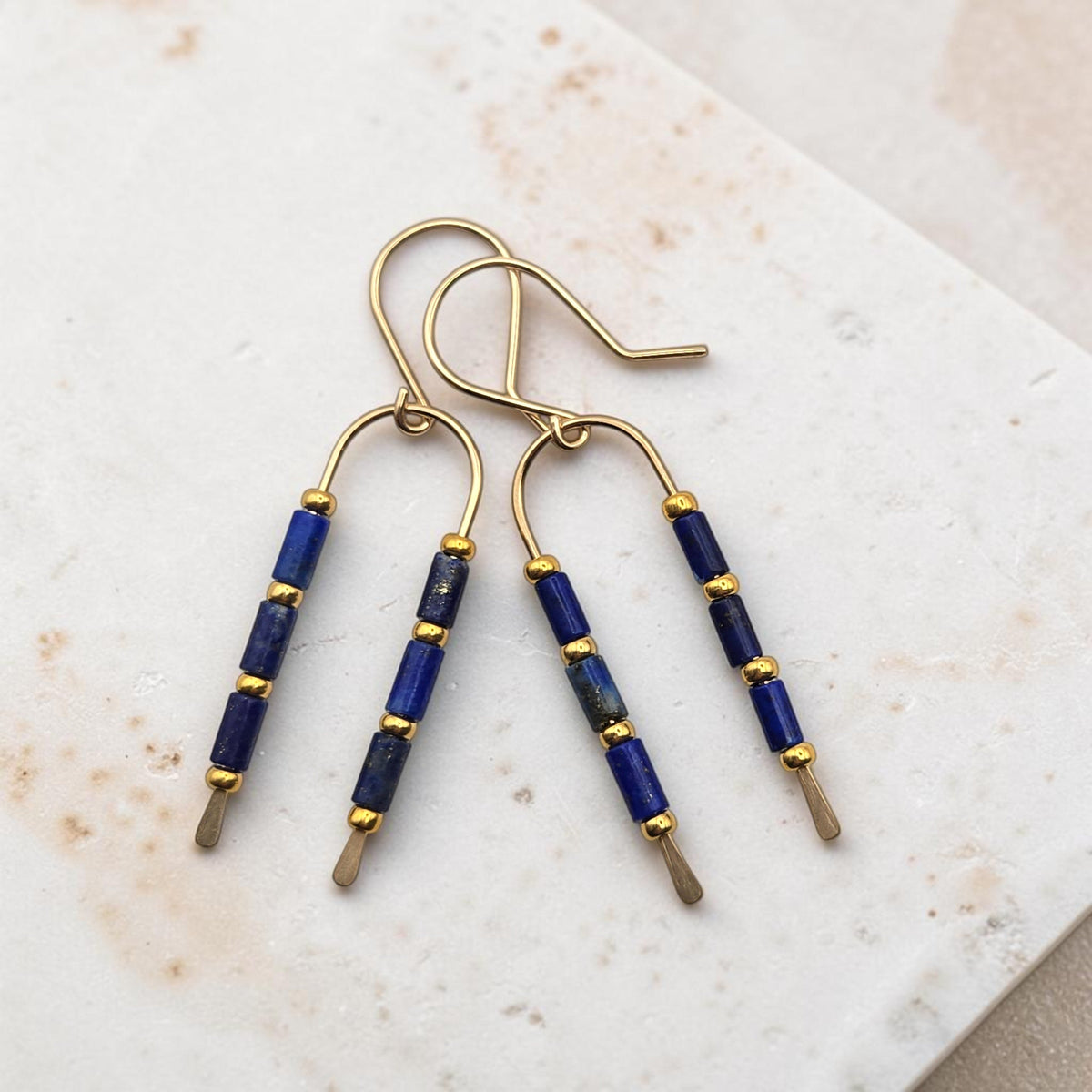 Bijou by SAM handcrafted gold wishbone earrings featuring blue Lapis Lazuli tube stones on a marble surface.