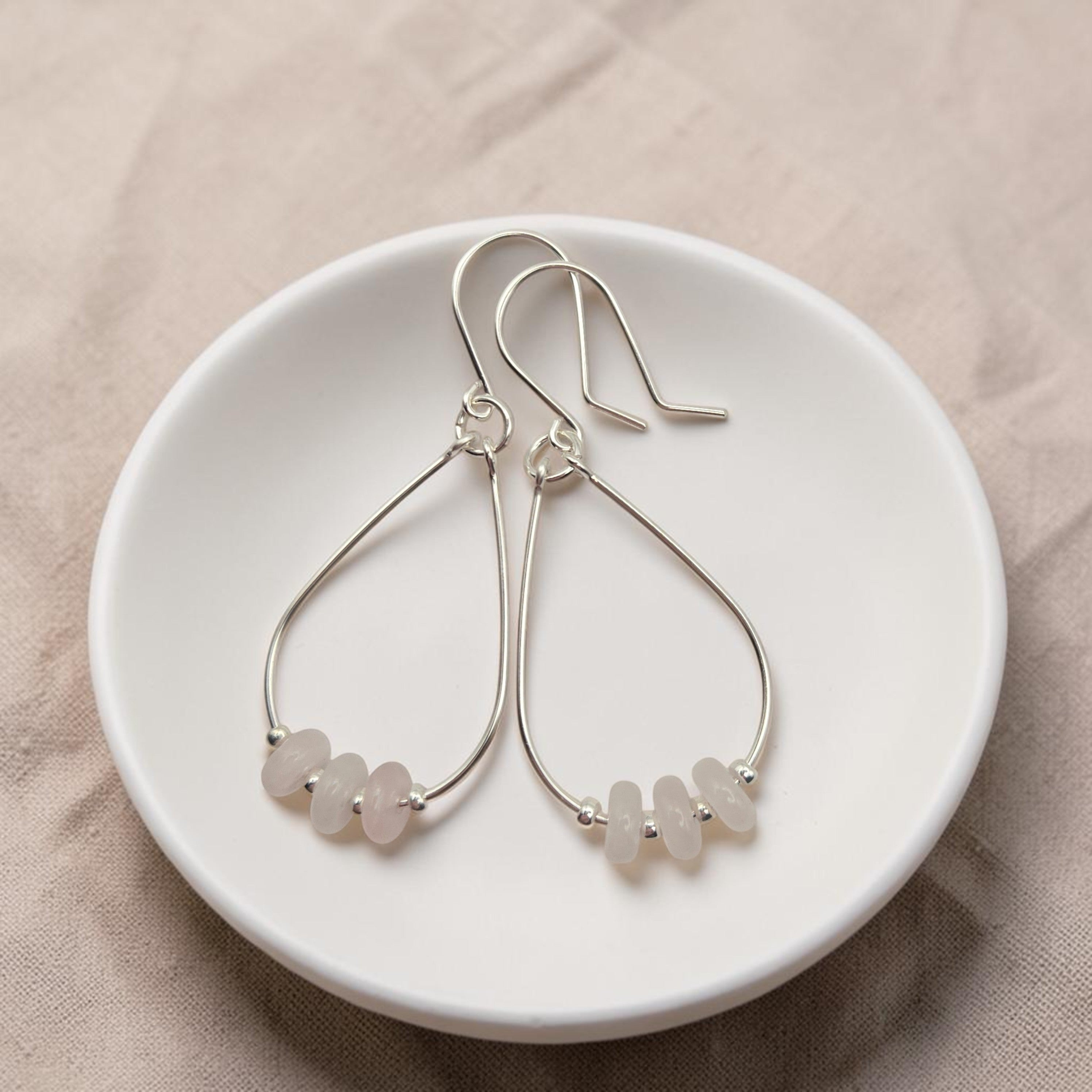 Bijou by SAM Aurora teardrop hoop earrings featuring blush rose quartz beads on sterling silver wire in a white dish.